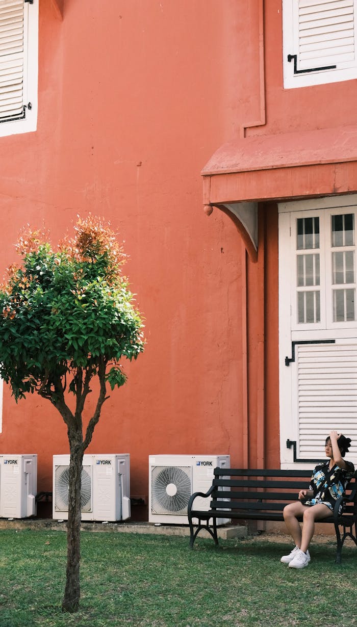 A woman relaxes on a bench by a red building with air conditioners.